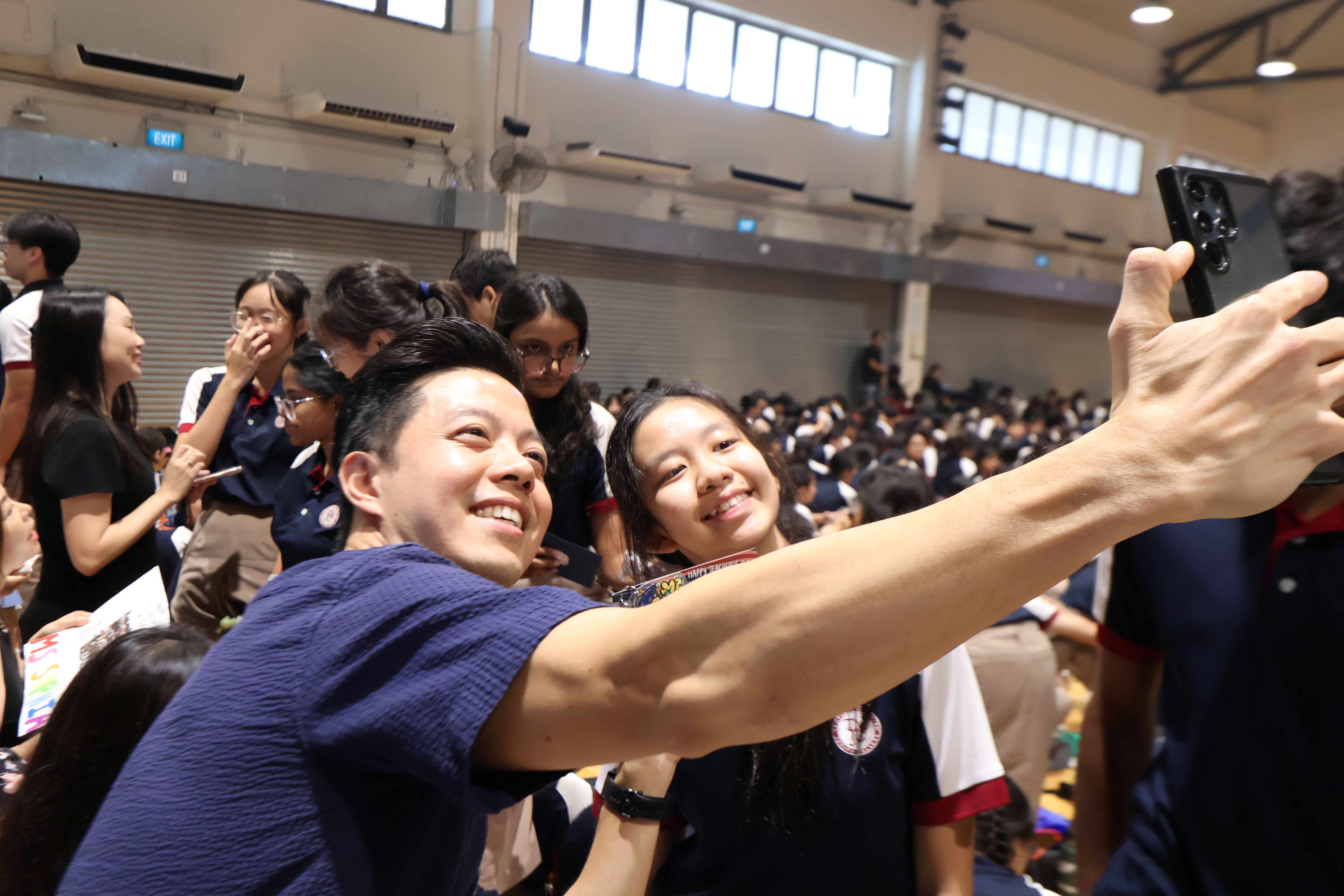 A teacher and a student lean in together, smiling broadly as they take a selfie in a packed school hall filled with students in the background.